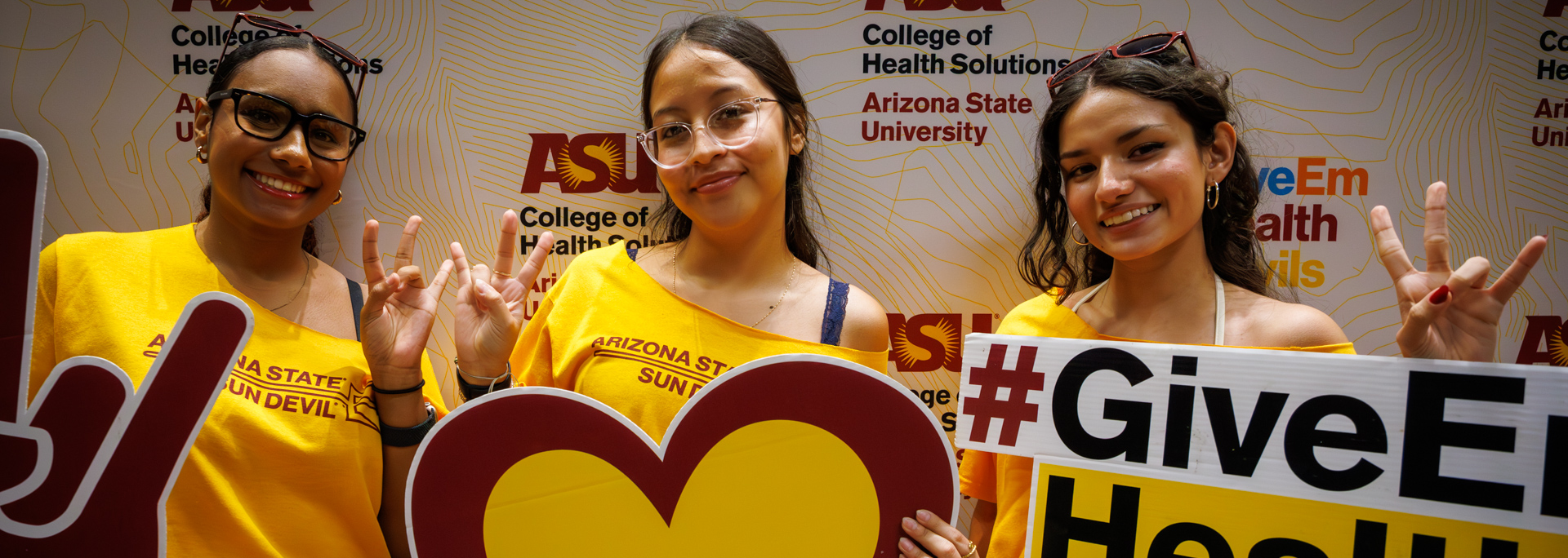 Three ASU students in gold shirts pose with signs and pitchfork hand signs in front of a College of Health Solutions backdrop, showing pride and spirit during a campus celebration.