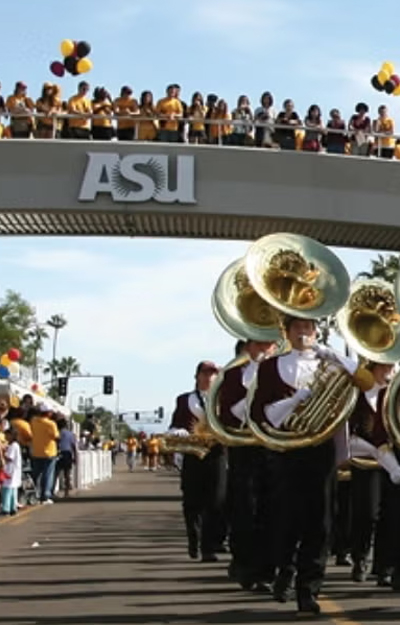 A marching band performs in an ASU homecoming parade, passing under a pedestrian bridge crowded with cheering spectators dressed in gold and maroon.